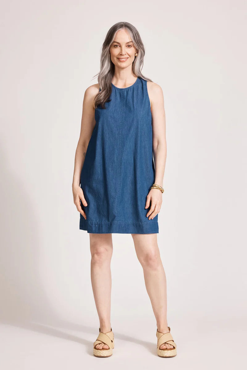 An indigo chambray tank dress made from lightweight 100% cotton, featuring exposed shoulders and a slight A-line silhouette. The dress includes contrast topstitching, a button closure at the back neck, a wide cuffed hem, and functional pockets for added practicality. Its breezy, denim-inspired look offers an effortless, summery style