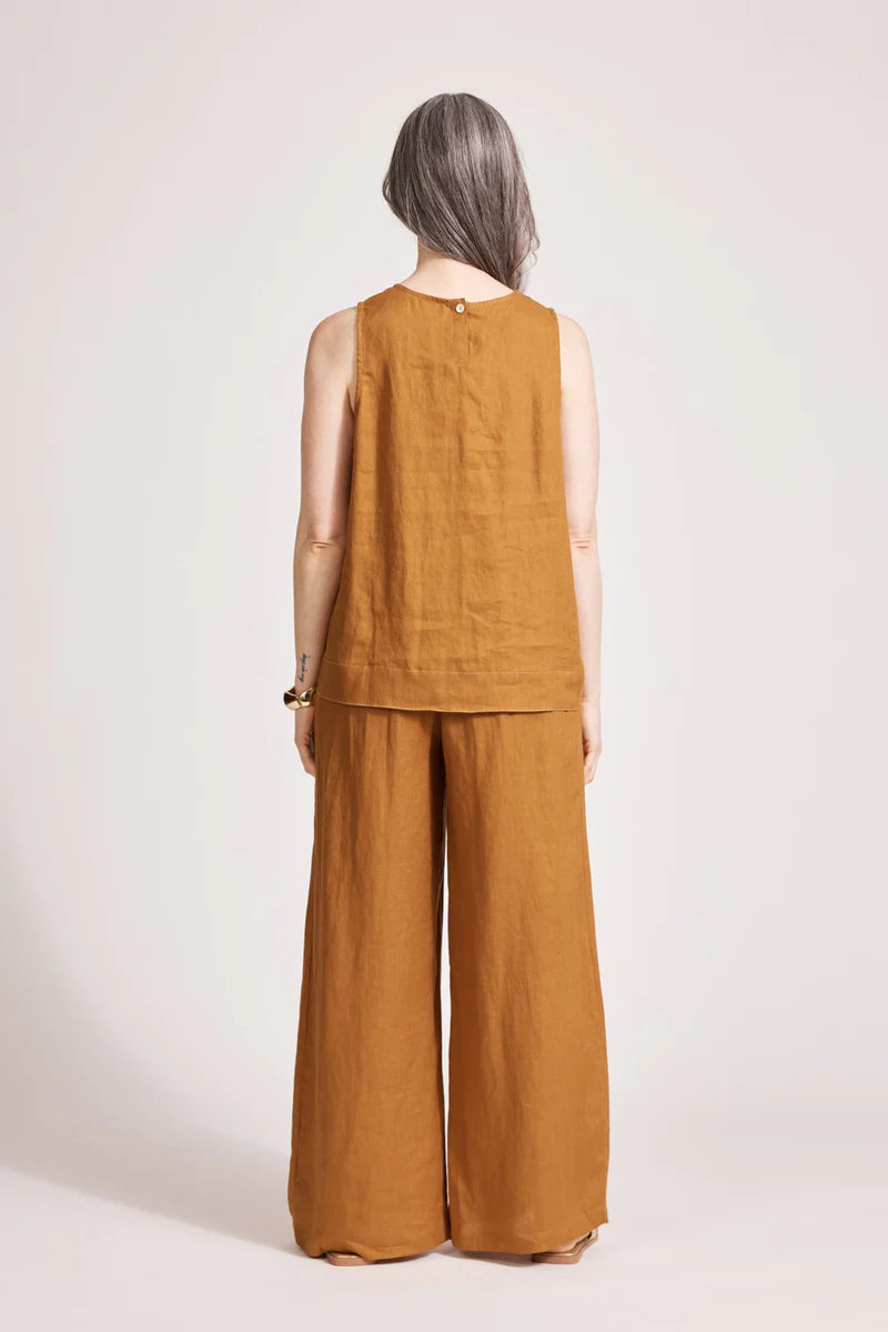 A relaxed, caramel-coloured 100% linen tank top featuring exposed shoulders, contrast decorative topstitching, and a soft A-line silhouette. The design includes a wide cuffed hem and a button closure at the back neck, giving the lightweight, breathable top an effortlessly refined look