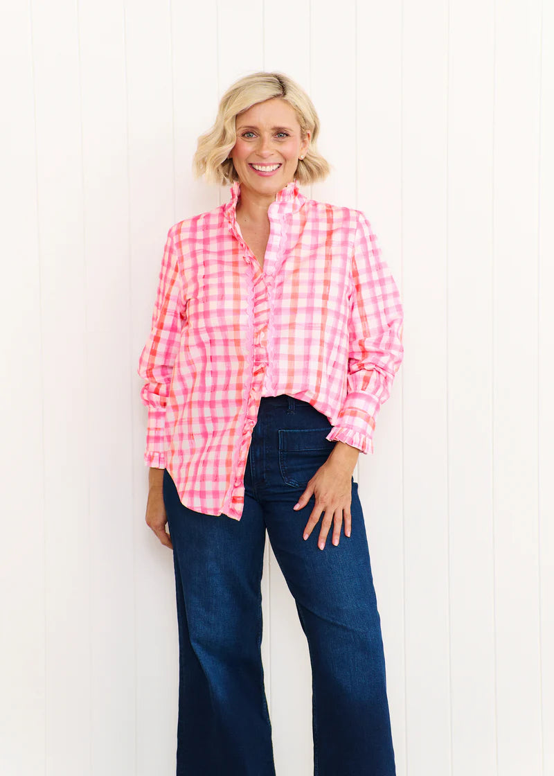 Woman wearing the Tilly Shirt in pastel pink and orange print, featuring a ruffled stand collar, button front, and lightweight cotton fabric, styled for a relaxed beachside look.