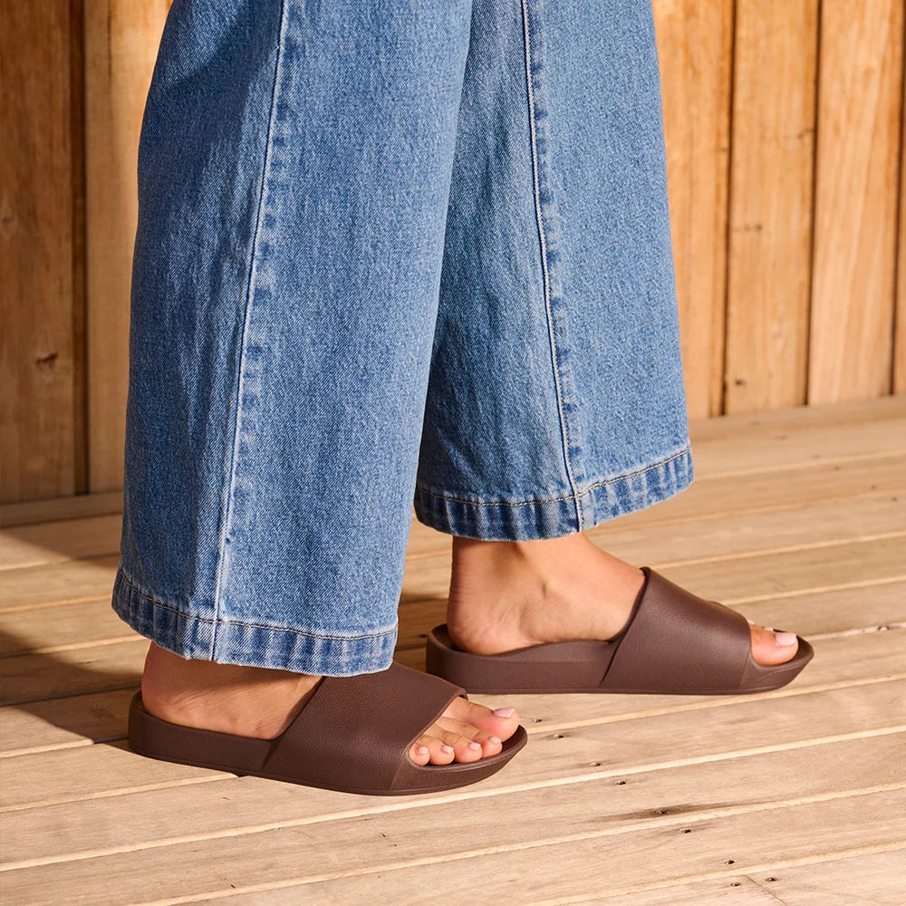 A pair of chocolate brown arch support slides with contoured footbeds and wide straps, designed for comfort and all-day wear.