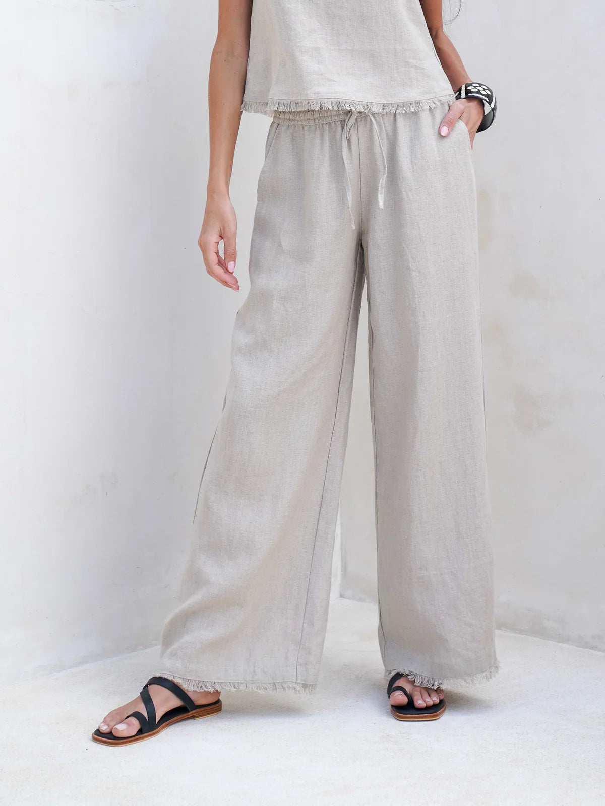 Natural French linen coastal pants with a relaxed fit, elastic waistband, and frayed-edge hem, styled for an effortless summer look.
