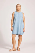 A denim chambray tank dress made from lightweight 100% cotton, featuring exposed shoulders and a slight A-line silhouette. The dress includes contrast topstitching, a button closure at the back neck, a wide cuffed hem, and functional pockets for added practicality. Its breezy, denim-inspired look offers an effortless, summery style