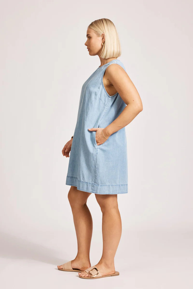 A denim chambray tank dress made from lightweight 100% cotton, featuring exposed shoulders and a slight A-line silhouette. The dress includes contrast topstitching, a button closure at the back neck, a wide cuffed hem, and functional pockets for added practicality. Its breezy, denim-inspired look offers an effortless, summery style