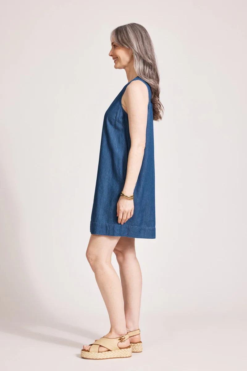 An indigo chambray tank dress made from lightweight 100% cotton, featuring exposed shoulders and a slight A-line silhouette. The dress includes contrast topstitching, a button closure at the back neck, a wide cuffed hem, and functional pockets for added practicality. Its breezy, denim-inspired look offers an effortless, summery style