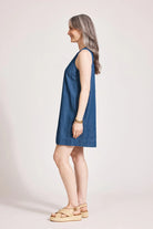 An indigo chambray tank dress made from lightweight 100% cotton, featuring exposed shoulders and a slight A-line silhouette. The dress includes contrast topstitching, a button closure at the back neck, a wide cuffed hem, and functional pockets for added practicality. Its breezy, denim-inspired look offers an effortless, summery style