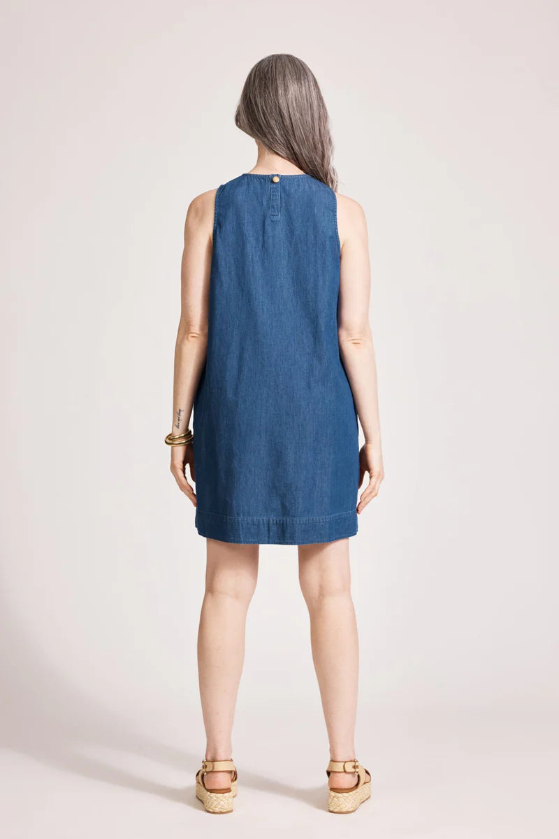 An indigo chambray tank dress made from lightweight 100% cotton, featuring exposed shoulders and a slight A-line silhouette. The dress includes contrast topstitching, a button closure at the back neck, a wide cuffed hem, and functional pockets for added practicality. Its breezy, denim-inspired look offers an effortless, summery style