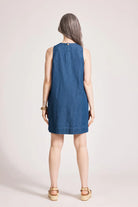 An indigo chambray tank dress made from lightweight 100% cotton, featuring exposed shoulders and a slight A-line silhouette. The dress includes contrast topstitching, a button closure at the back neck, a wide cuffed hem, and functional pockets for added practicality. Its breezy, denim-inspired look offers an effortless, summery style
