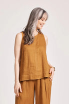 A relaxed, caramel-coloured 100% linen tank top featuring exposed shoulders, contrast decorative topstitching, and a soft A-line silhouette. The design includes a wide cuffed hem and a button closure at the back neck, giving the lightweight, breathable top an effortlessly refined look
