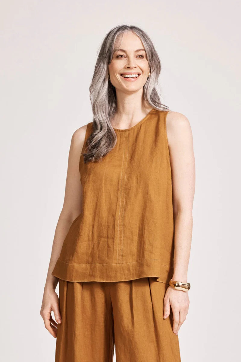 A relaxed, caramel-coloured 100% linen tank top featuring exposed shoulders, contrast decorative topstitching, and a soft A-line silhouette. The design includes a wide cuffed hem and a button closure at the back neck, giving the lightweight, breathable top an effortlessly refined look