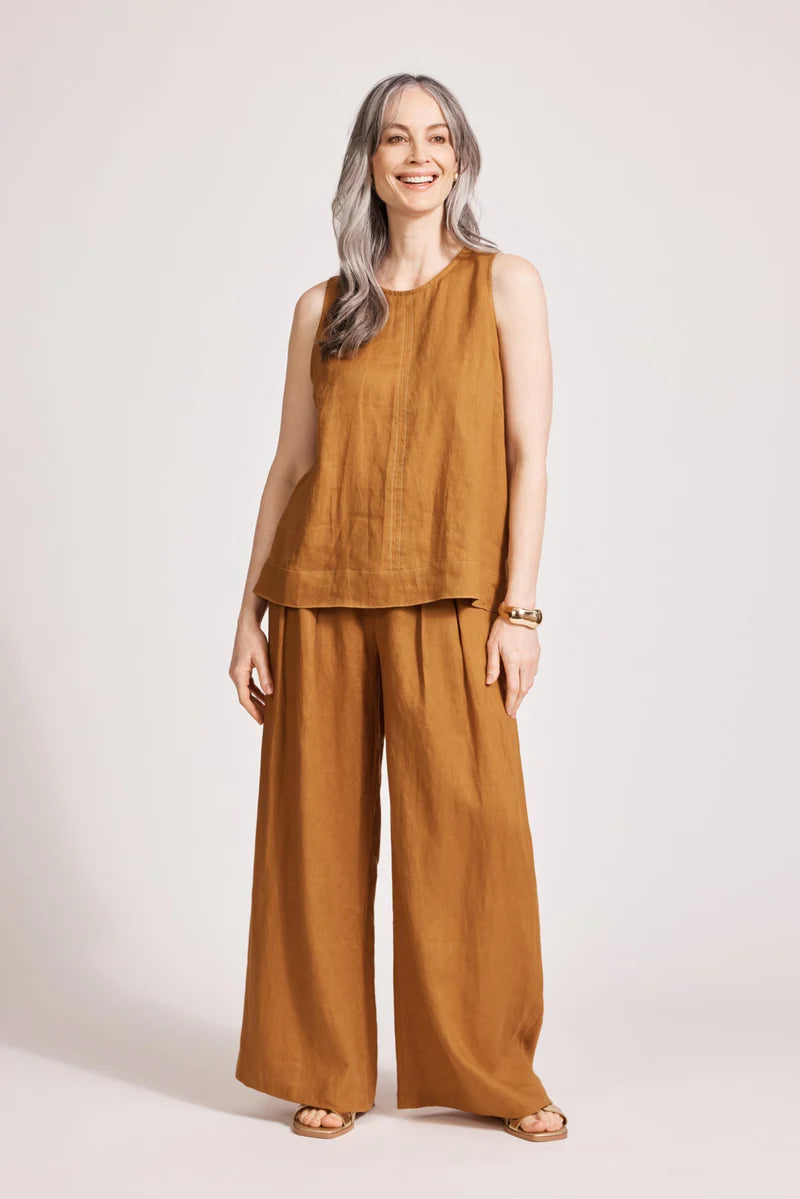 A relaxed, caramel-coloured 100% linen tank top featuring exposed shoulders, contrast decorative topstitching, and a soft A-line silhouette. The design includes a wide cuffed hem and a button closure at the back neck, giving the lightweight, breathable top an effortlessly refined look