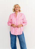 Woman wearing the Tilly Shirt in pastel pink and orange print, featuring a ruffled stand collar, button front, and lightweight cotton fabric, styled for a relaxed beachside look.
